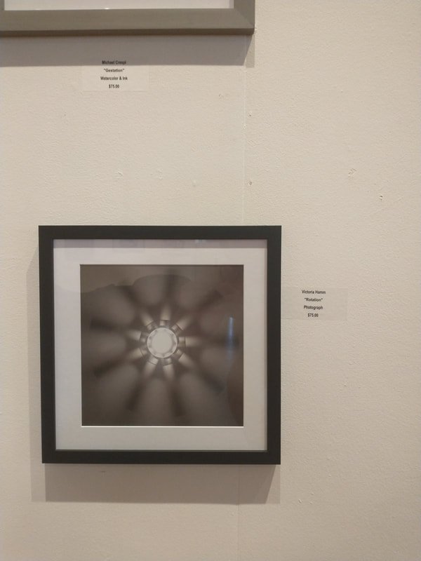 A framed black-and-white photograph of a spiral staircase viewed from above, hung on a white gallery wall with price tags indicating it is titled 'Rotation' by Victoria Horne, priced at $75.00.