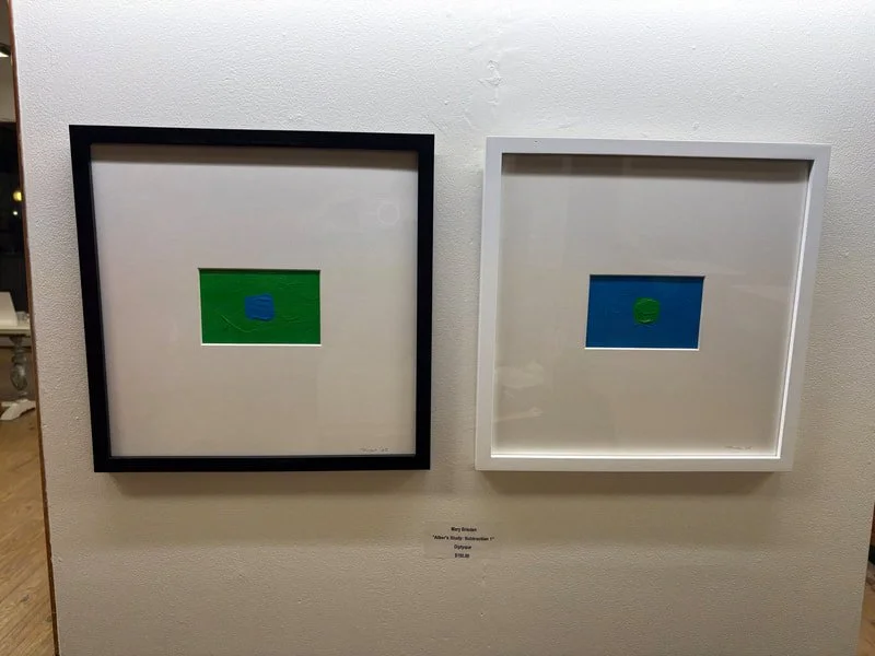Two framed minimalist artworks on a white wall, each with a central circle on a colored background—one with a green background and a blue circle, the other with a blue background and a green circle.