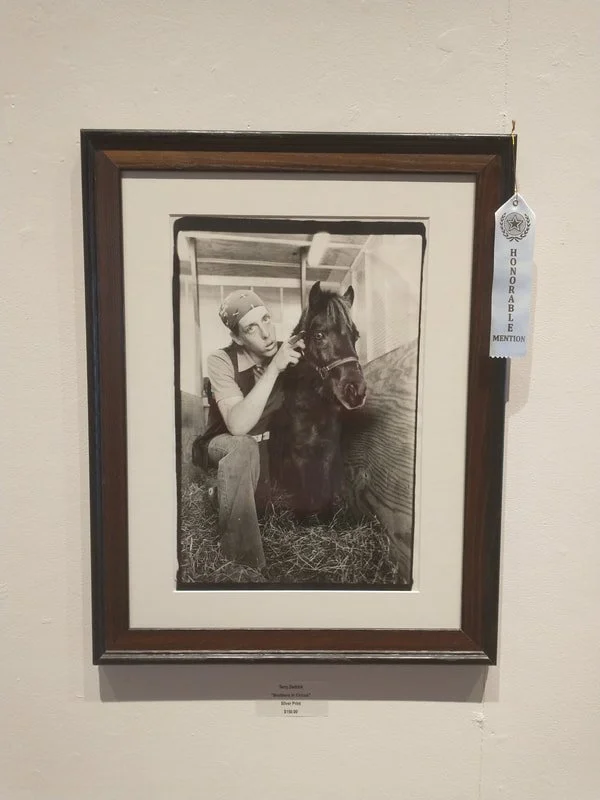 Black and white photograph of a woman grooming a small horse, in a stable or barn setting.