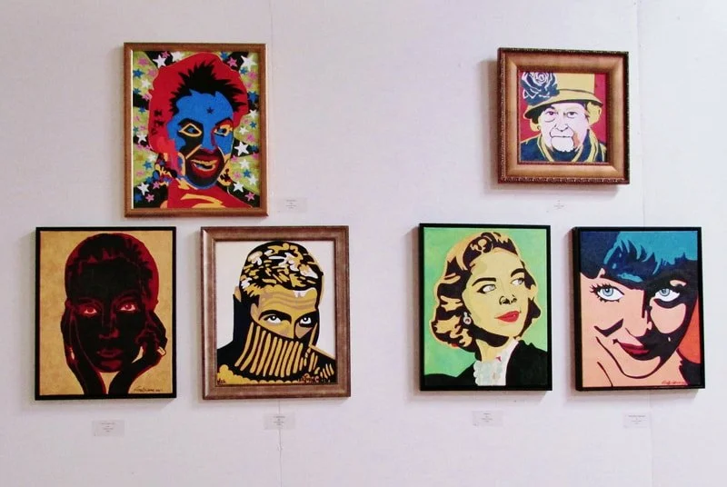 Six colorful pop art portraits displayed on a white gallery wall. The portraits feature various faces with vibrant backgrounds and bold outlines.