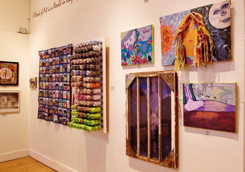 Gallery wall with colorful textile art, mixed media paintings, and a sculptural portrait of a woman with long, multicolored yarn hair.