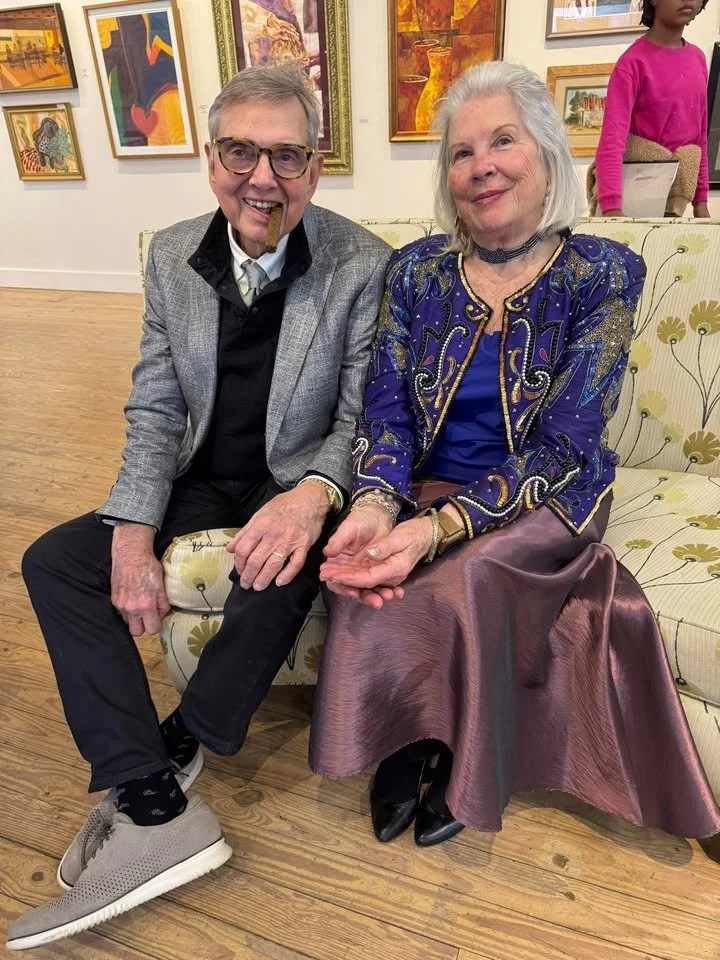 Two elderly people sitting on a cream-colored sofa with green and yellow floral pattern, smiling. The man on the left has gray hair, glasses, a cigar in his mouth, a gray blazer, black shirt, black pants, and gray sneakers. The woman on the right has