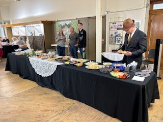 A buffet table with various dishes and desserts at an indoor event, with people serving themselves and socializing in the background.