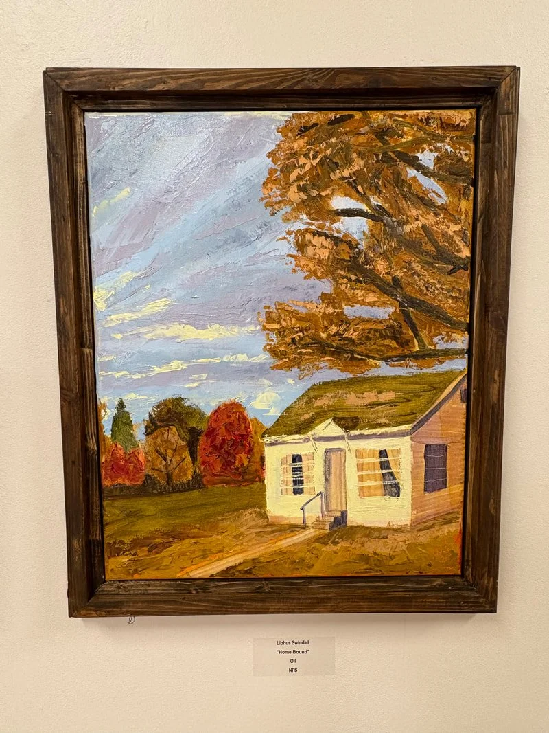 An oil painting of a small house with a green roof, surrounded by autumn trees with colorful foliage. The scene includes a cloudy sky and a dirt path leading to the house, framed in dark wood.