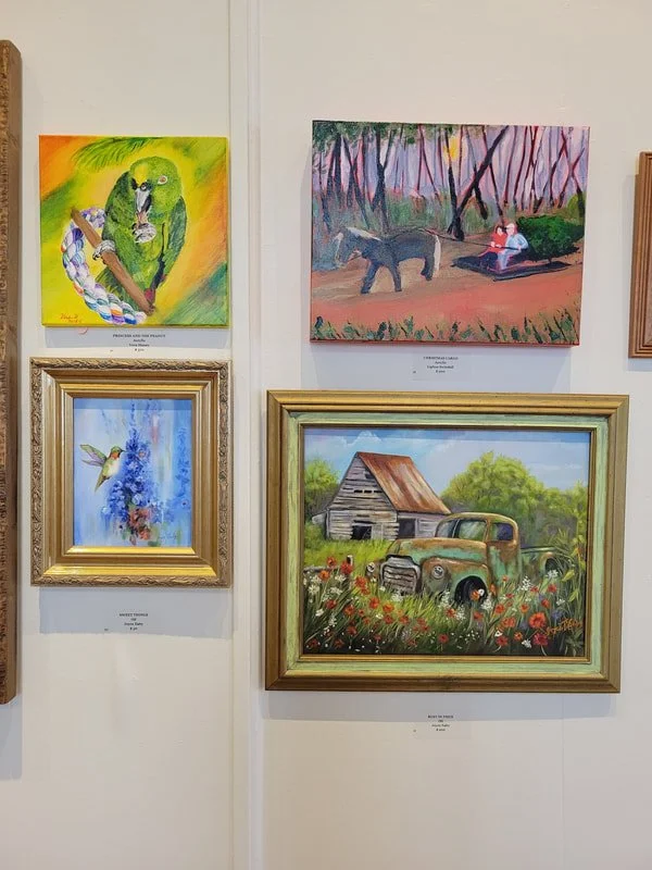 A white wall displaying four paintings, including a colorful lion with a rainbow mane, a girl riding an elephant in a forest, a hummingbird near flowers, and an old truck beside a barn in a field of flowers.