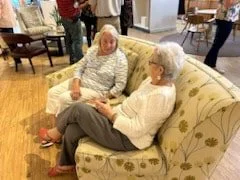 Two elderly women sitting and talking on a floral-patterned sofa in a room with wooden flooring and other people in the background.