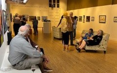 People gathered and sitting in an art gallery, viewing paintings on the walls.