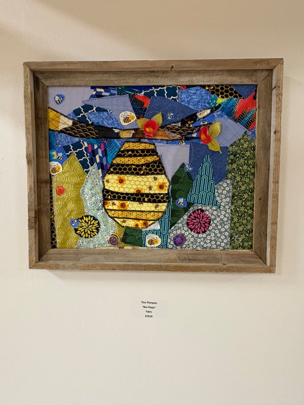 Fabric art depicting a bee with a yellow and black striped body hanging on a wall, framed in wood, with a label underneath.
