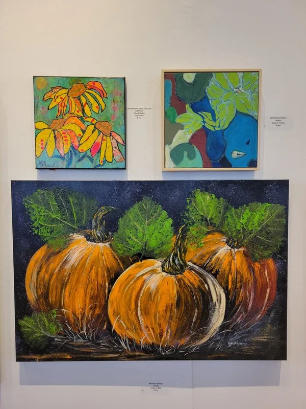 Three painted artworks on a gallery wall; two smaller abstract paintings above and one large pumpkin painting below featuring orange pumpkins and green leaves.