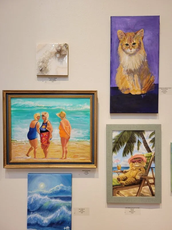 Art gallery wall displaying five paintings: a textured abstract piece, a portrait of a ginger cat with a purple background, three women in swimsuits walking on the beach, a scenic ocean wave at sunset, and a lounging cat in a tropical setting.