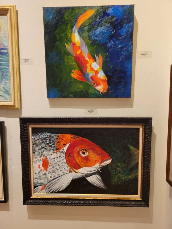 Two colorful fish paintings hung on a wall, one above the other. The top painting depicts an orange and white koi fish swimming in blue and green water. The bottom painting shows a large, red and white fish with black spots swimming in dark water wit
