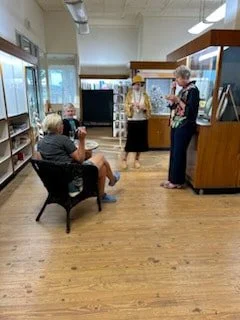 Four elderly people in a room, two sitting on chairs and two standing near display cases, engaged in conversation.