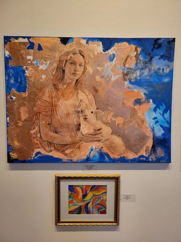 Painting of a woman with long hair holding a small white puppy, with a background of abstract ashen gold and blue colors, and a smaller colorful abstract painting with swirling patterns framed below.