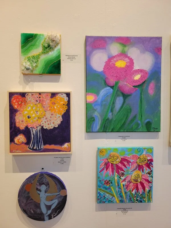 An art exhibit with five paintings and one decorative plate on display. The top left is a green abstract piece with textured elements. The top right features a floral painting of pink flowers with yellow centers against a purple and blue background. 