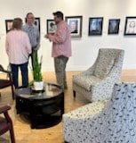 Three people having a conversation in a room with framed pictures on the wall, a large potted plant, and a patterned armchair.