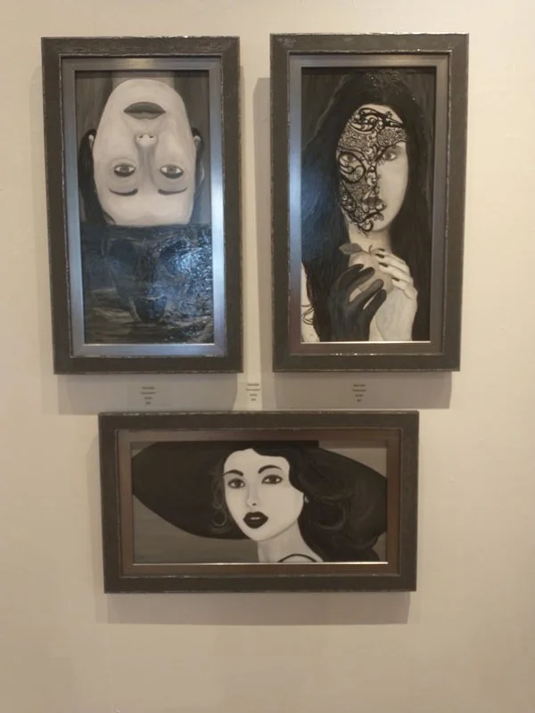 Three black and white portrait paintings of women displayed on a wall. The top-left portrait is upside down, featuring a woman with long dark hair and a calm expression. The top-right portrait shows a woman with long dark hair, partially covered by a