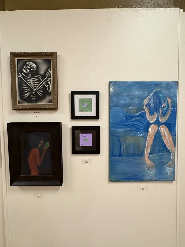 Art gallery wall with six framed paintings including a skeleton, an abstract portrait, and a person sitting on a bed with their head bowed.
