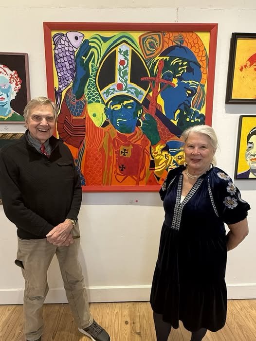 Two people, a man and a woman, standing in front of colorful, modern-style artwork at an art gallery. The man on the left is wearing a black jacket over a gray shirt, beige pants, and glasses. The woman on the right is wearing a black dress with flor