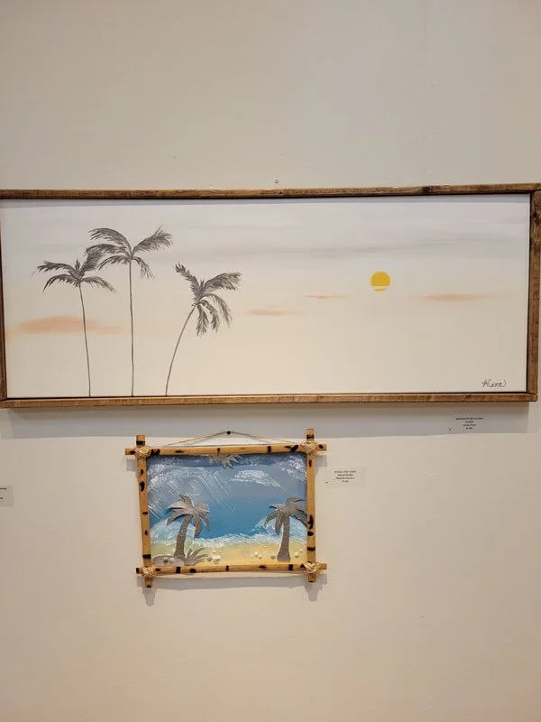 Two paintings of tropical beach scenes with palm trees, one large and one small, displayed on a wall in an art gallery.