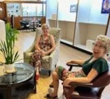 Two women sitting and smiling in a cozy room with plants and a window.
