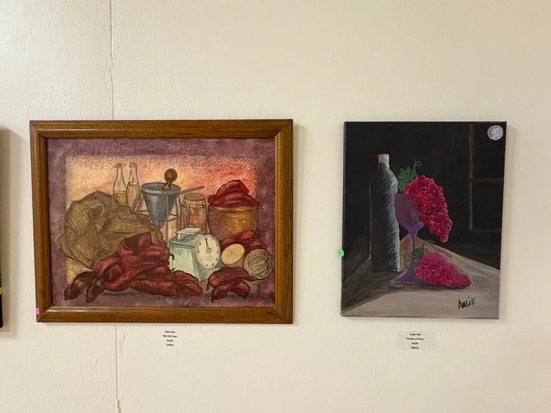 Two still life paintings hung on a white wall. The left one depicts a variety of food items including bread, a clock, bottles, and meats. The right one shows a wine bottle, a bunch of grapes, and a purple wine glass on a table with a dark background.