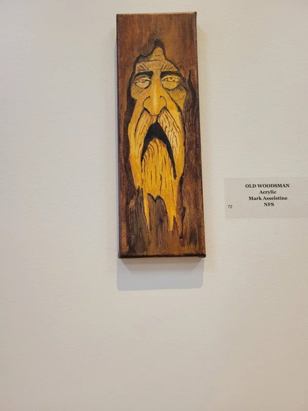 Wooden plaque with a painted portrait of an old woodsman with a long yellow beard and mustache, and a stern expression, displayed on a white wall.