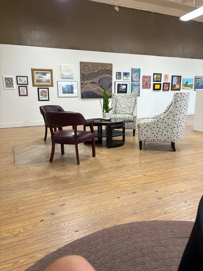 An art gallery with a seating area consisting of three chairs around a small black round table with a potted plant. The background displays various framed artworks on a white wall, and the floor is wooden.