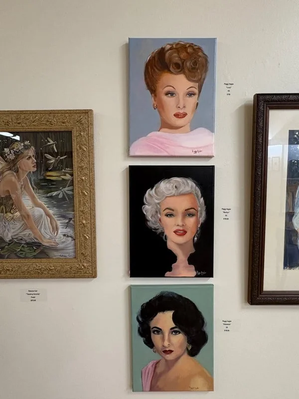 Three paintings of women hung on a wall, with another artwork partially visible on the left. The top painting shows a woman with short curly reddish hair, the middle shows a woman with grey curly hair, and the bottom depicts a woman with short dark c