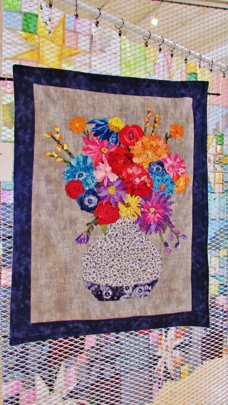 Colorful embroidered quilt with a flower bouquet in a vase design hanging on a display net.