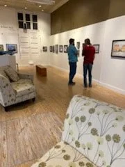 Two people viewing artwork in an art gallery with white walls and wooden flooring, with a sofa in the foreground.