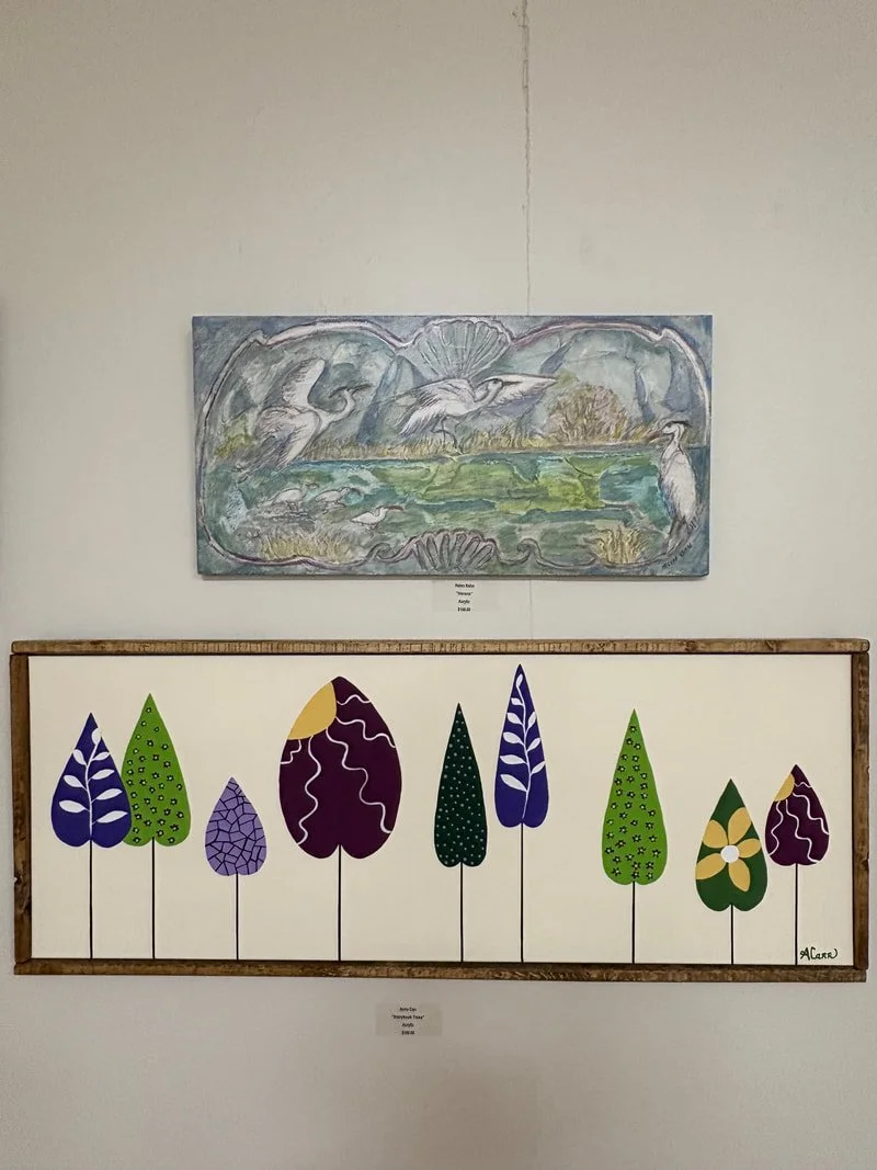 A colorful abstract painting of herons above a landscape, and a framed artwork of stylized, colorful trees on a white wall.