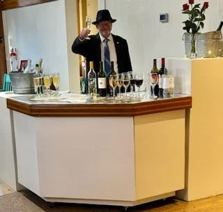 A man in a suit and hat standing behind a bar with various bottles of alcohol and glasses, raising one hand in a toast.