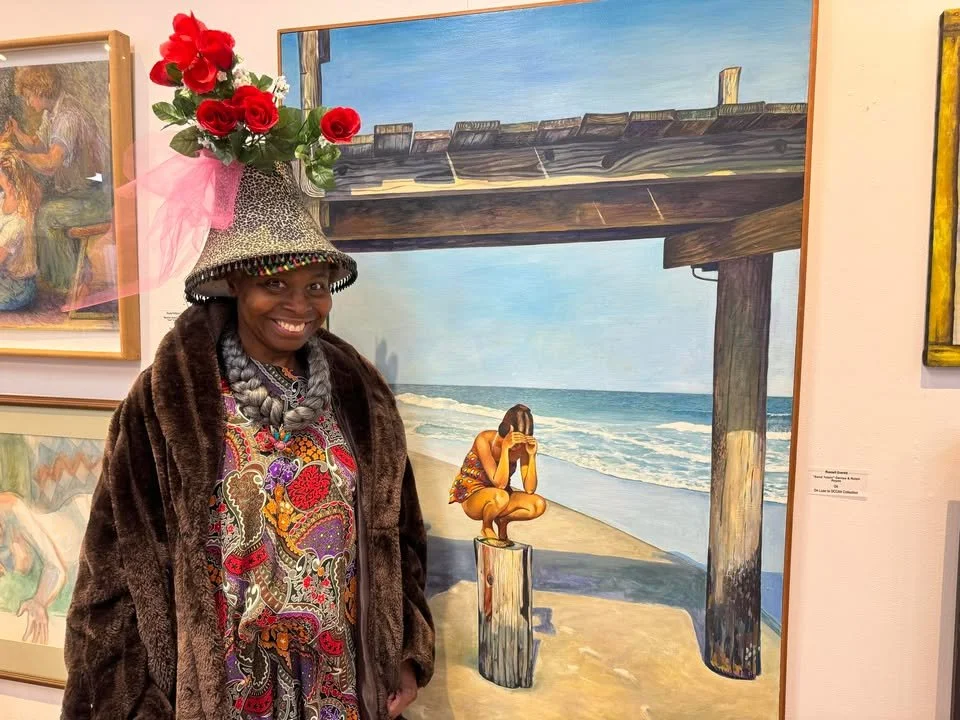 Woman in a patterned dress and fur coat wearing a hat decorated with red roses and pink tulle standing in front of a beach painting at an art gallery.