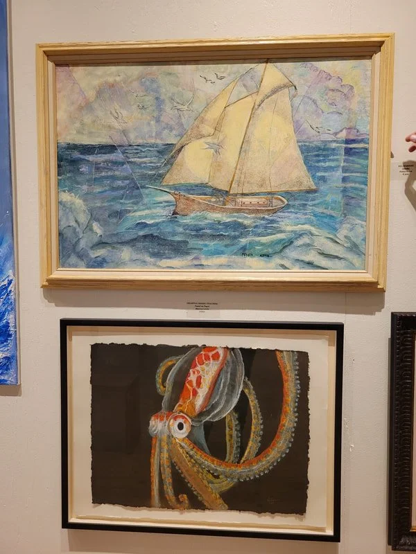 A painting of a sailing ship on the ocean with cloudy sky and seagulls, framed in light wood. Below it, a painting of an octopus with orange and white pattern on a dark background, framed in black.