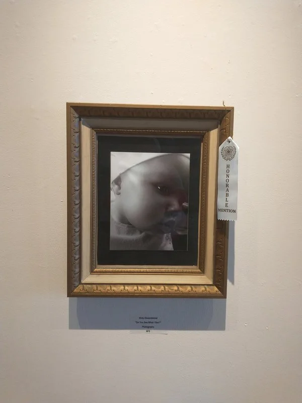Photograph of a baby with a pacifier in their mouth, framed and displayed on a wall, with an 'Honorable Mention' ribbon attached beside it.