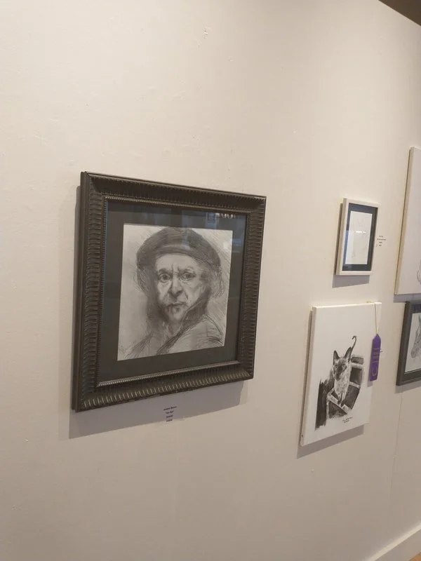 A framed charcoal sketch of an older woman with glasses and a headscarf hanging on a white gallery wall, alongside other artworks.