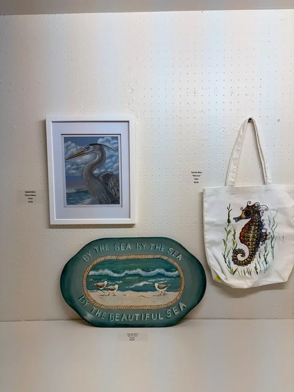 A framed painting of a heron standing in water with clouds in the sky in the background, a decorative ceramic piece with sea waves and seagulls, and a white tote bag with a colorful seahorse design hanging on a white wall.