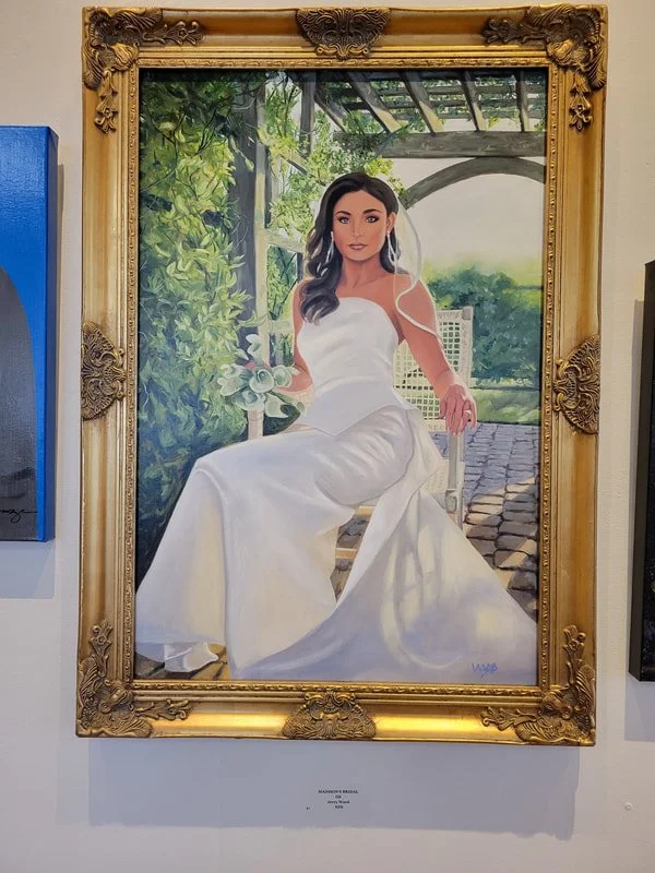 Painting of a woman in a white wedding dress sitting outdoors on a white chair, holding a bouquet, with greenery and a stone path in the background, framed by an ornate gold frame.
