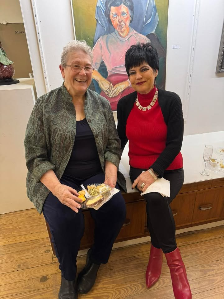 Two women sitting together at an art gallery or exhibition, with a colorful portrait painting of an older woman in the background. One woman is holding a plate of food, and there are empty glasses on the table next to them.