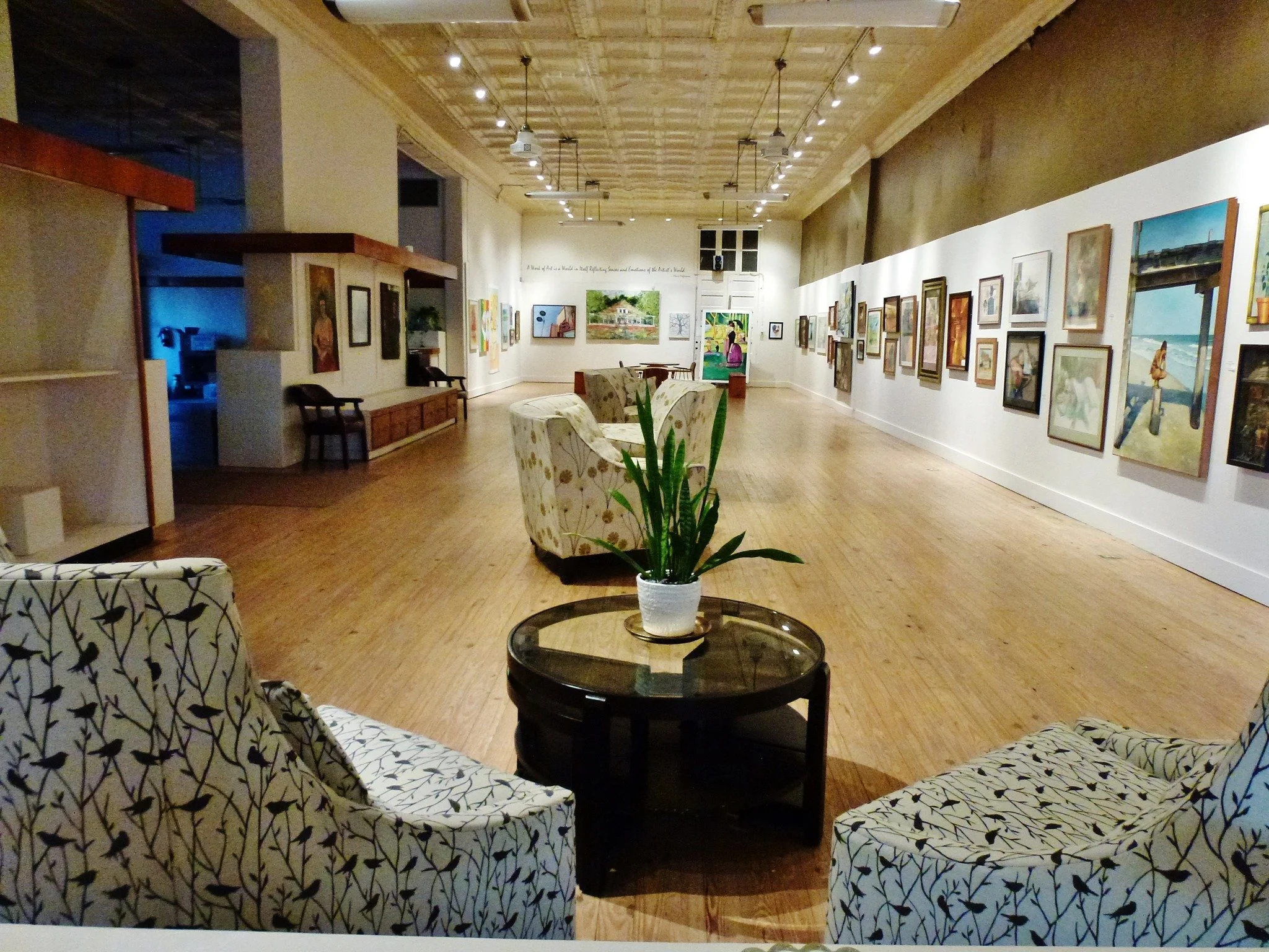 An art gallery with framed artwork on the walls, a seating area with patterned armchairs, a console table, and a large potted plant in the center, illuminated by wall and ceiling lights.