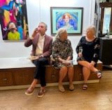 Three elderly women sitting on a wooden bench, looking at their phones inside an art gallery with framed artwork on the wall behind them.
