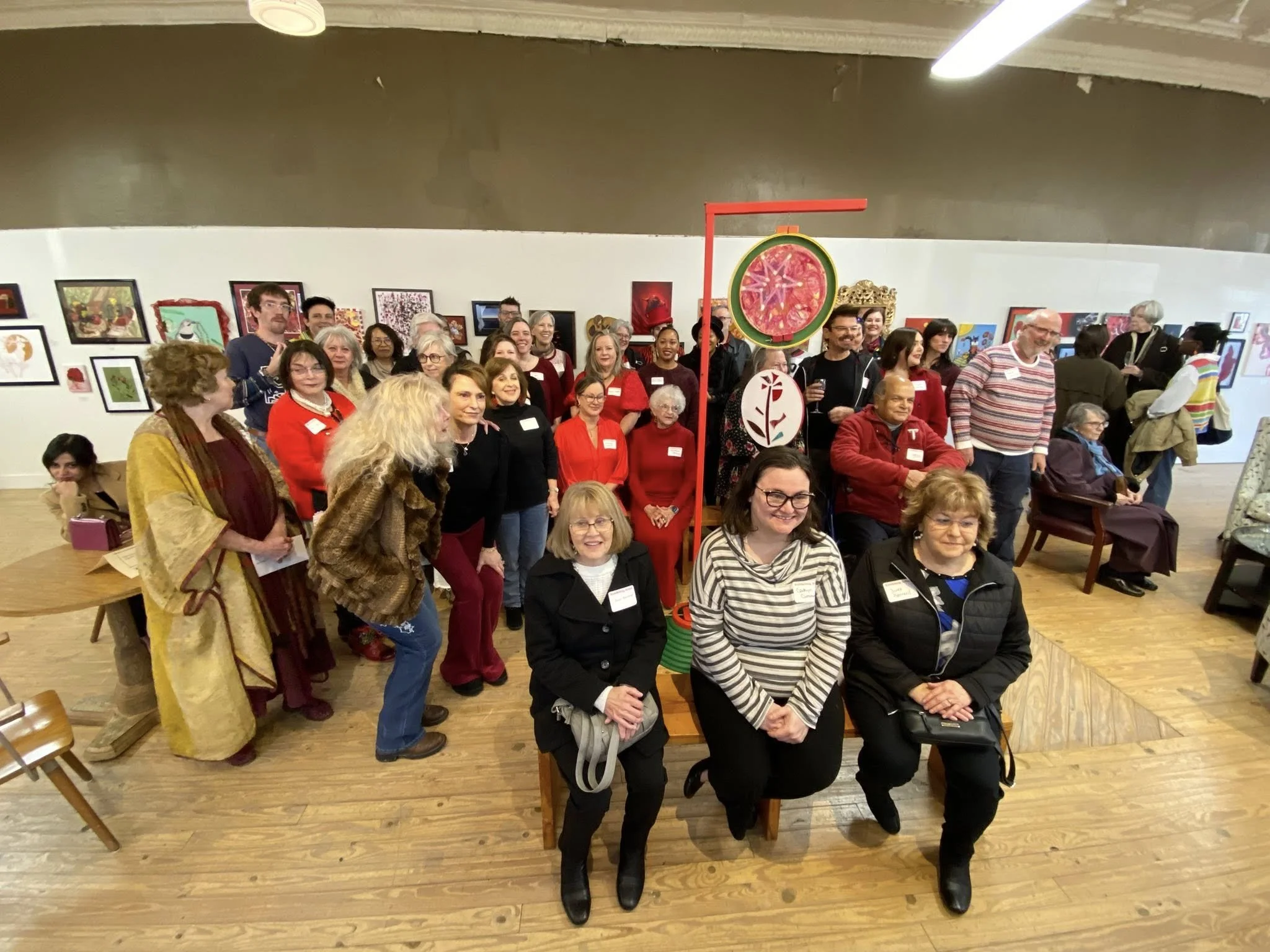 A large group of people gathered in an art gallery, some standing and some seated, surrounded by framed artwork on the walls. The group appears diverse, with many smiling, and some wearing name tags. There are decorative art pieces on stands, including a large fake pizza slice, and wooden flooring. The atmosphere looks lively and social.