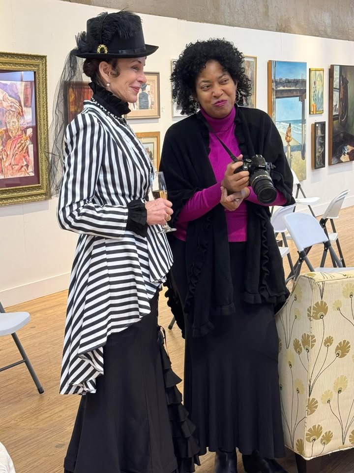 Two women standing in an art gallery, one dressed in a Victorian-style striped dress with a hat and holding a champagne flute, the other in a purple turtleneck with a camera, surrounded by framed artwork on the wall.