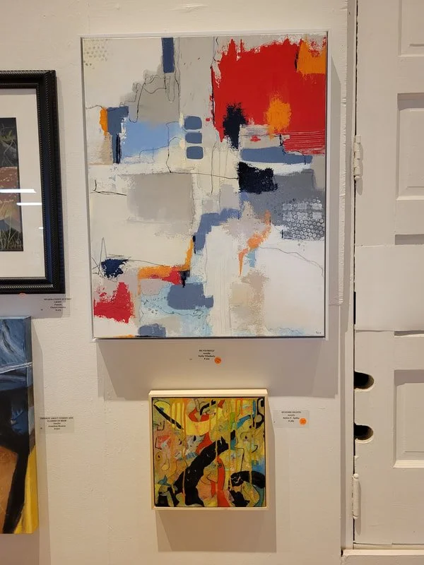 Two abstract paintings displayed on a white gallery wall. The larger painting at the top features splashes of red, black, blue, gray, and beige with textured patterns. The smaller painting below has bright yellow, red, black, and green colors with sw