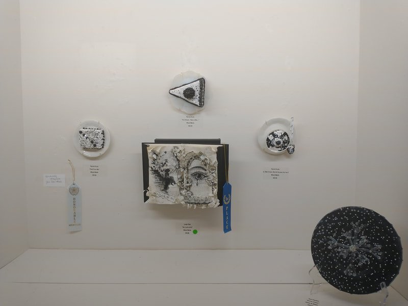 Art display featuring five ceramic pieces, a fabric art piece, and a decorated umbrella on a white wall and surface.
