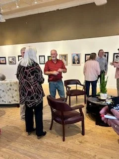 People viewing artwork in an art gallery with framed photos on the wall, a plant, and chairs.