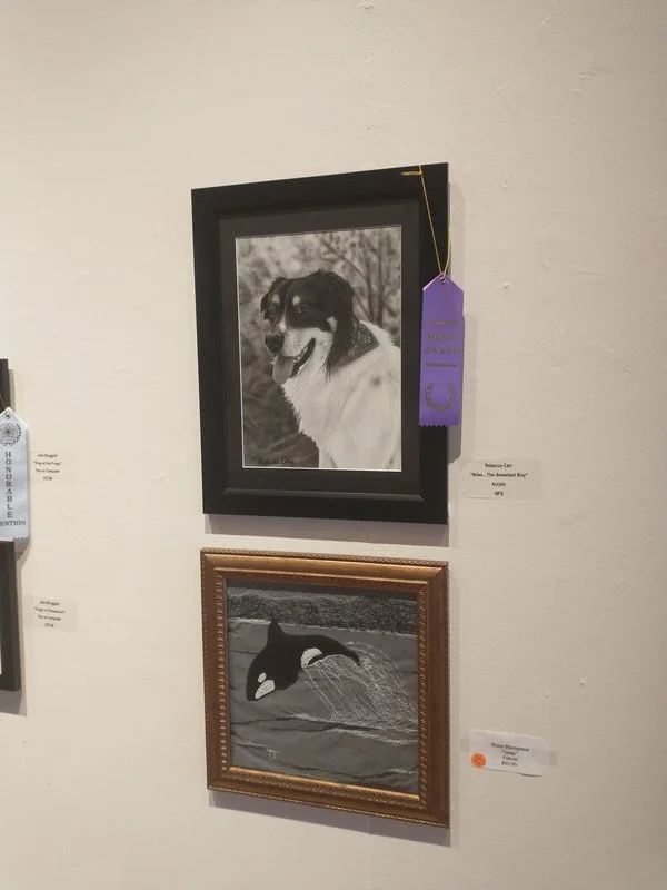 Two framed photographs on a white wall, the top one is a black and white photo of a dog with a happy expression, and the bottom one is an artwork of an orca whale in water.