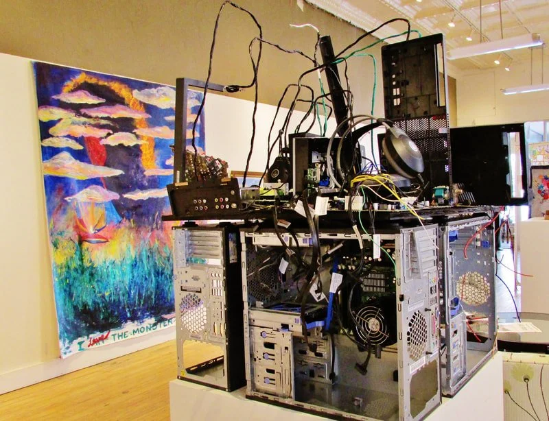An art installation featuring an open computer tower with wires and electronic components inside, placed on a white platform in front of a colorful abstract painting in a gallery space.