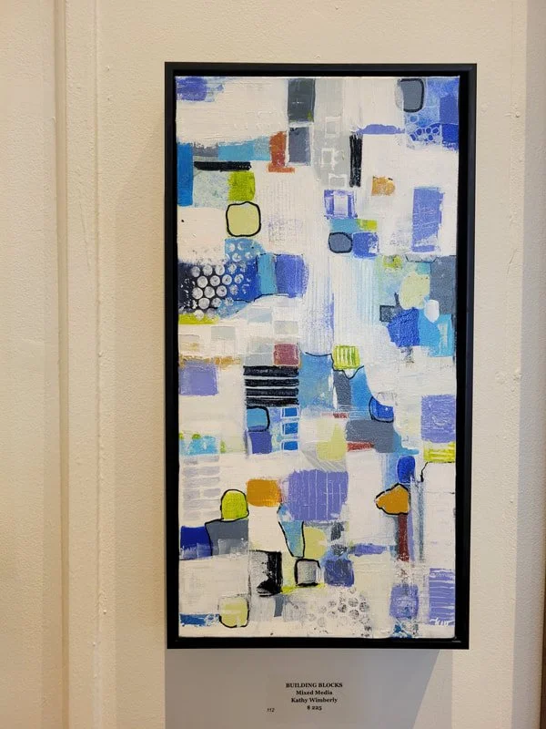 An abstract painting titled 'Building Blocks' with various geometric shapes in shades of blue, yellow, black, and gray on a white background, framed and displayed on a wall.