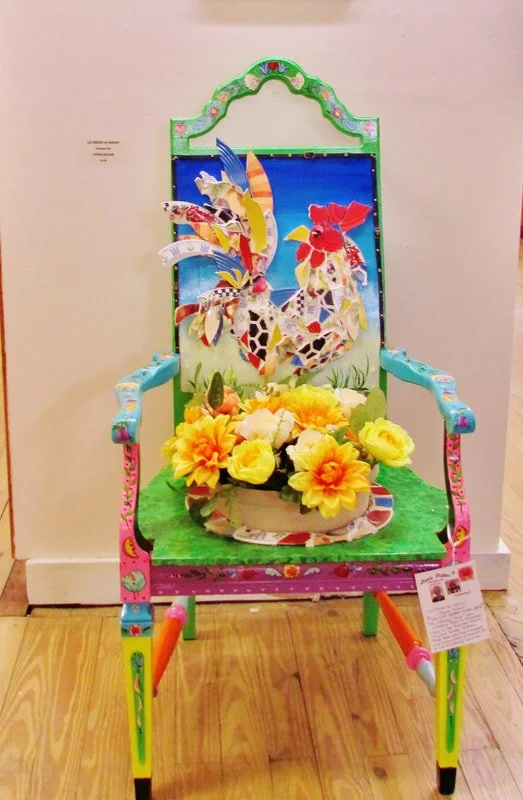 Colorful chair decorated with paper cutouts of roosters and a floral centerpiece with yellow and cream flowers.